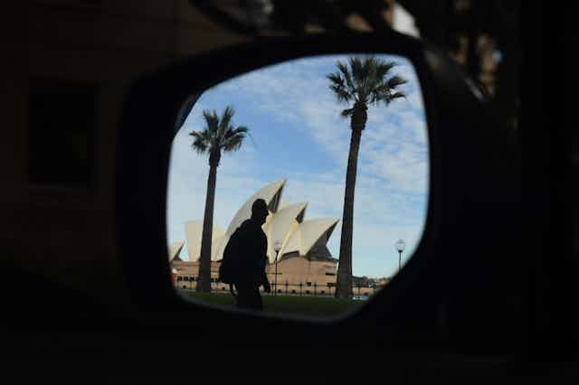 A man in front of the Sydney Opera House seen in a car side-view mirror