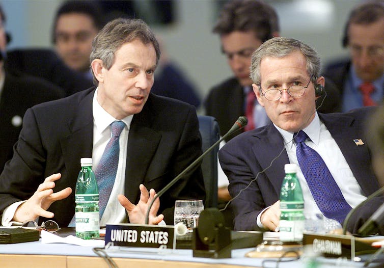 Tony Blair speaks and George W Bush listens, sat at table with microphones.