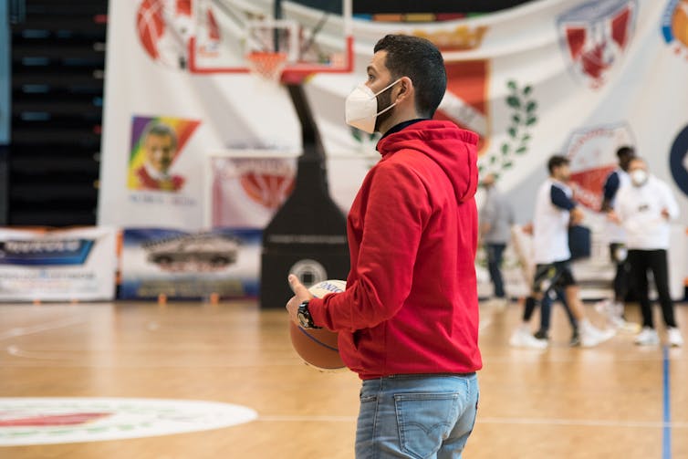 A man inside a recreation facility wears a mask and holds a basketball, he's on a basketball court and people are playing in the background