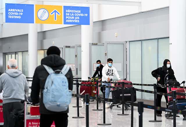 Travellers with luggage in an airport terminal under a sign directing them towards COVID-19 testing
