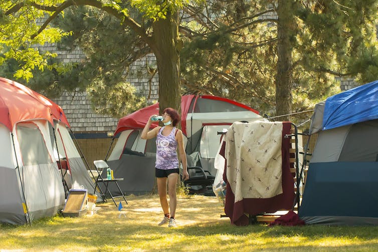 Woman drinks from plastic water bottle as she walks through pile of tents at homeless camp