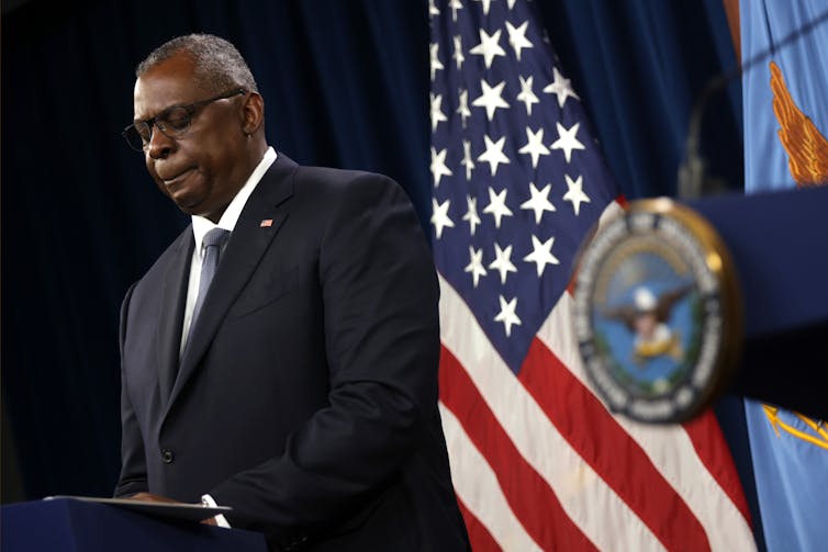 Defense Secretary Lloyd Austin standing at a lectern, lips pursed and eyes lowered, in front of an American flag.