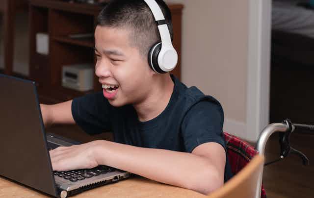 A boy in a wheelchair wearing headphones smiles at a laptop.