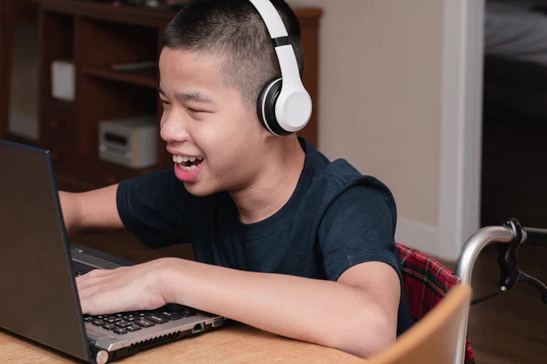 A boy in a wheelchair wearing headphones smiles at a laptop.