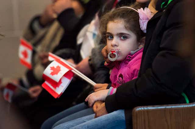 A young girl with a pacifier looks at the camera as others around her hold small Canadian flags.