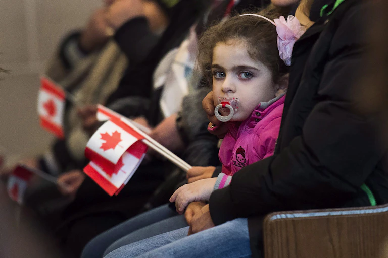 A young girl with a pacifier looks at the camera as others around her hold small Canadian flags.