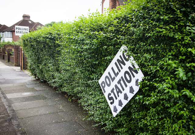 A sign for a polling station dangling precariously from a hedge. 