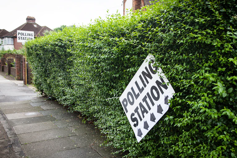 A sign for a polling station dangling precariously from a hedge.