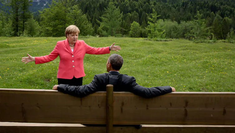 Angela Merkel stands in conversation with Barack Obama, who is sitting on a bench, looking relaxed.