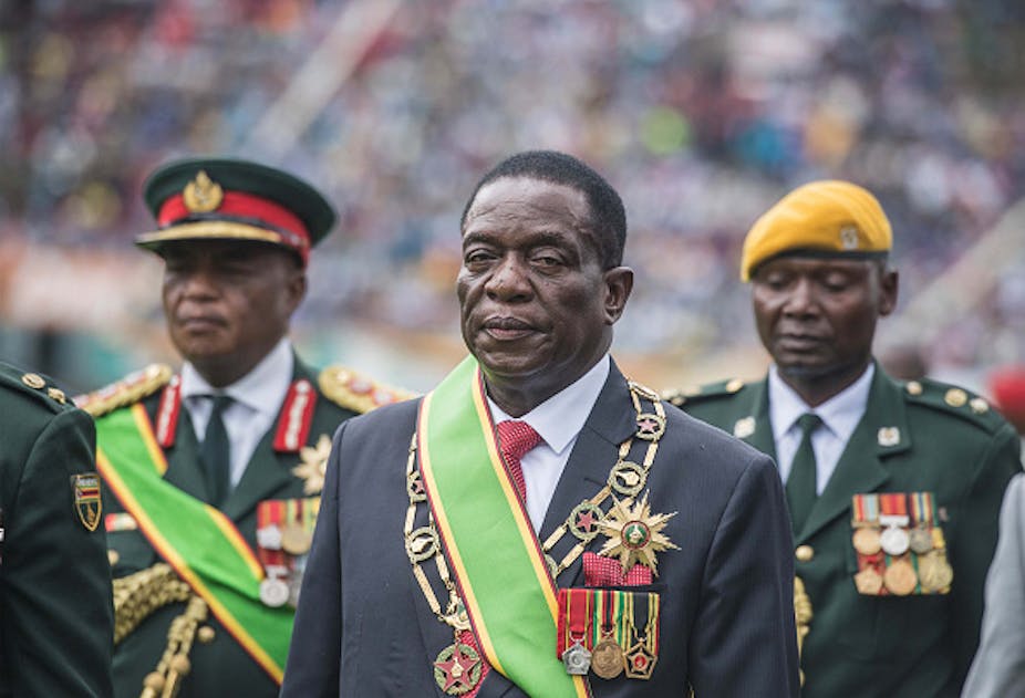 A man wearing a civilian suit decorated with military medals is flanked by two military officers. 