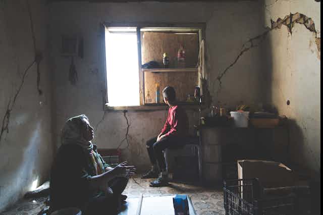 A woman and child sit in a boarded up room with a little light coming in through the window. There are cracks in the walls.