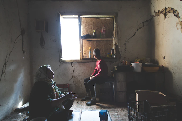 A woman and child sit in a boarded up room with a little light coming in through the window. There are cracks in the walls.