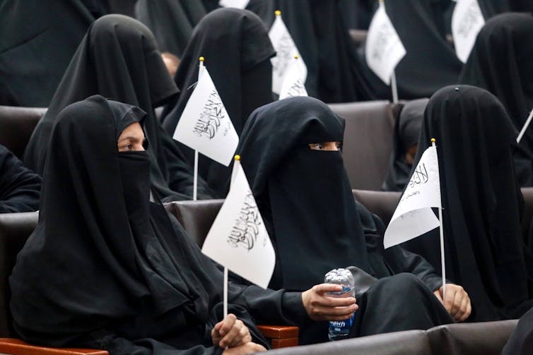 Rows of seated women wearing black niqabs and holding white Taliban flags.