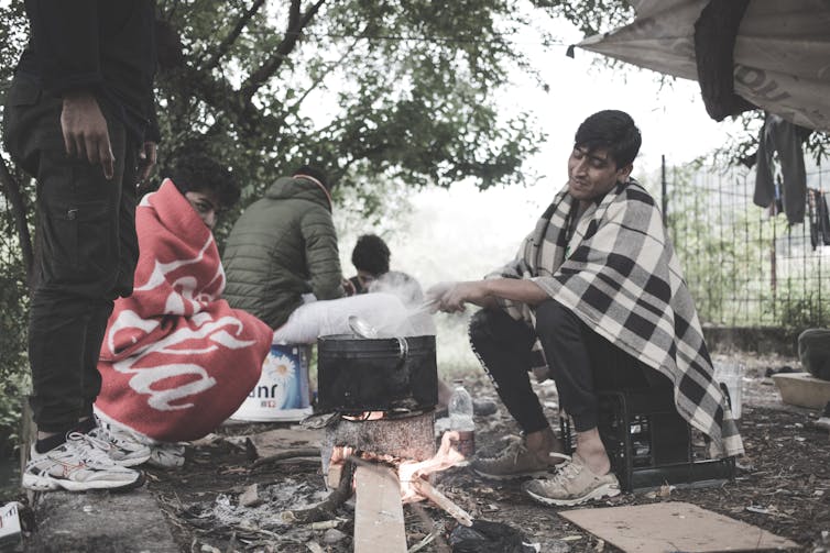Men wrapped in blankets sit around a pot boiling on a fire in a camp on the Bosnian border.