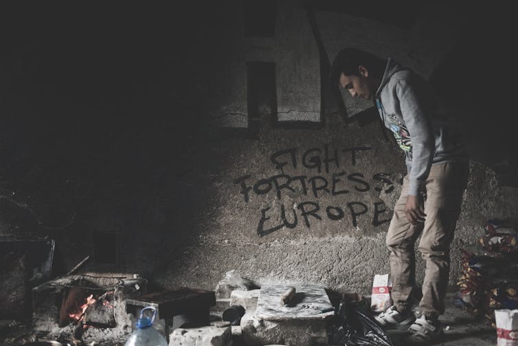 A man looks around a dark room in a migrant camp. Graffiti on the wall reads 'Fight Fortress Europe'.