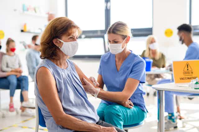 A woman in a face mask receives a vaccine from a woman in blue medical scrubs, also in a face mask