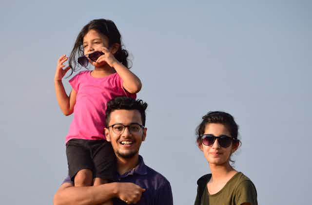 A girl in sunglasses stands on her father's shoulders, next to a mother.