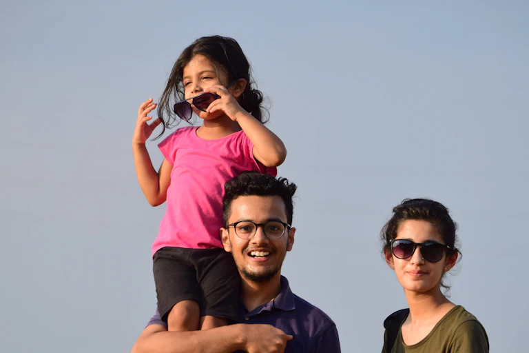 A girl in sunglasses stands on her father's shoulders, next to a mother.