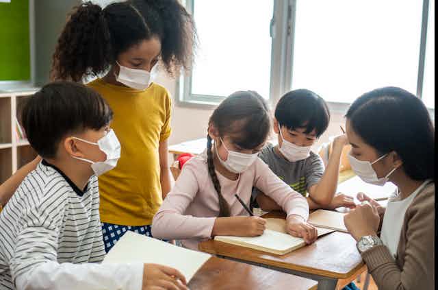 A teacher wearing a mask sits with her students at a desk.
