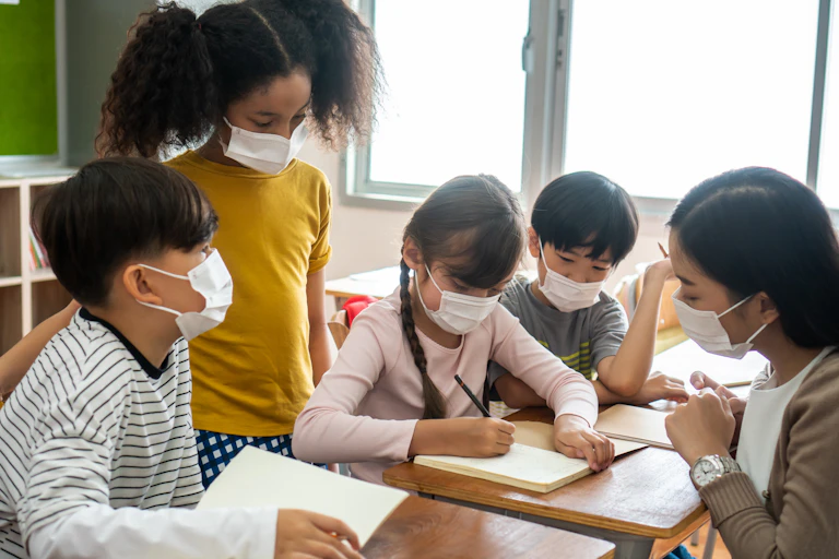 A teacher wearing a mask sits with her students at a desk.