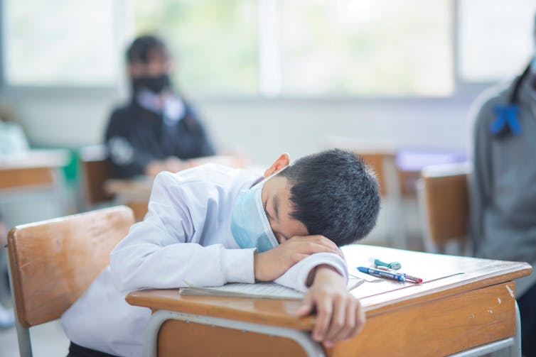 A youth in a face mask has his head laid down on a desk.