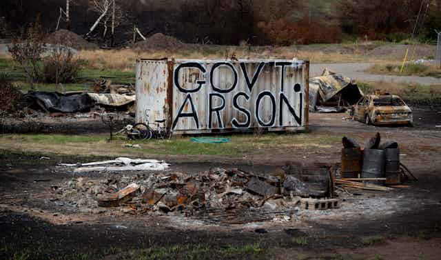 The words Government Arson are painted on a shipping container surrounded by burned debris.