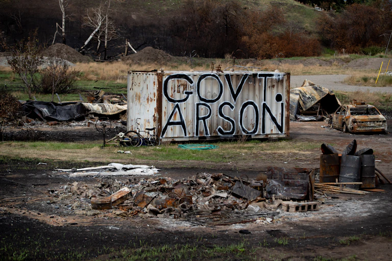 The words Government Arson are painted on a shipping container surrounded by burned debris.