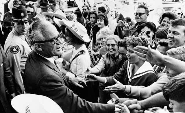 A black and white image of a man smiling and shaking hands with a crowd of people