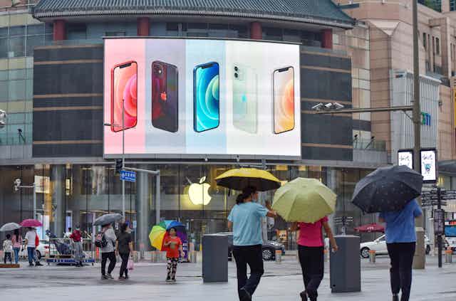 People carry umbrellas on a city street underneath security cameras while a large digital billboard in the background displays Apple iPhones