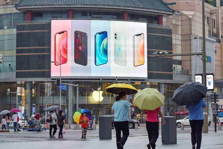 People carry umbrellas on a city street underneath security cameras while a large digital billboard in the background displays Apple iPhones