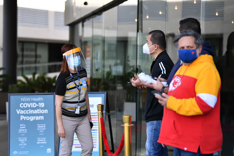 People in masks stand waiting outside a vaccination hub.