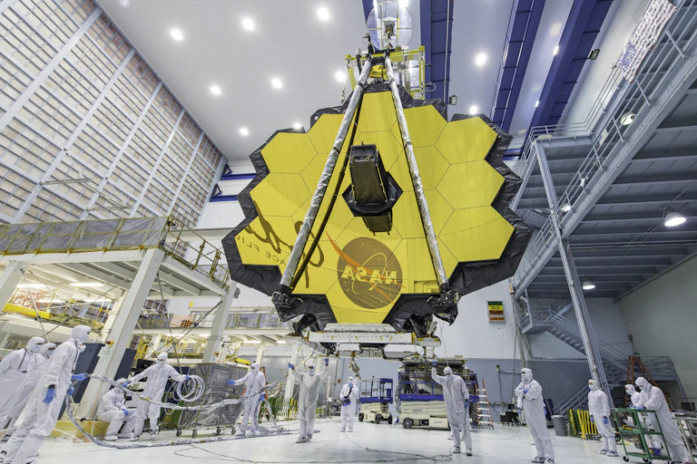 Workers in white coveralls standing below a large golden mirrored dish.