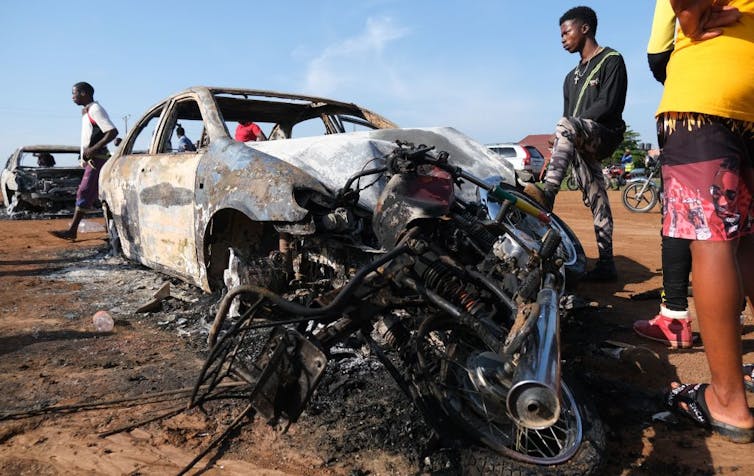 2 man standing beside the burnt remains of a car and some motorcycles.