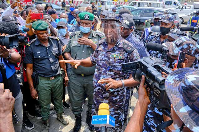 Men in police uniform surrounded by people pointing cameras and microphones at them. 
