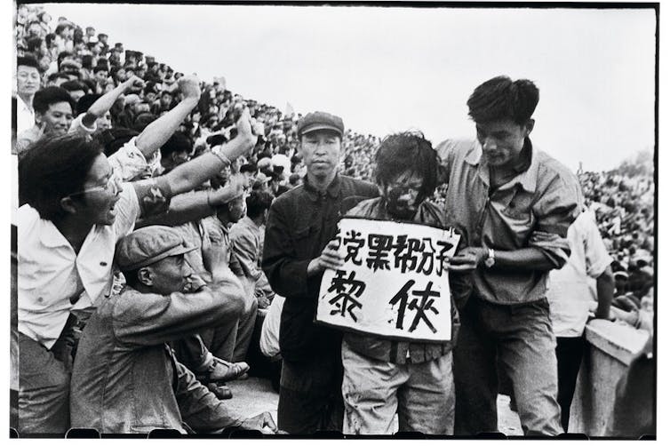 Black and white photo of Chinese man with a placard around his neck led through a jeering crowd throwing ink in his face.