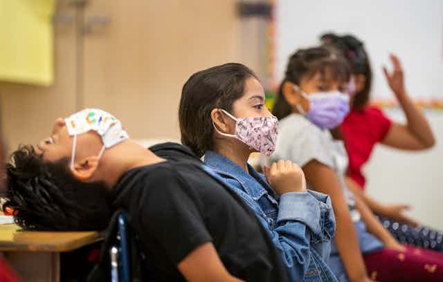 Four third-grade students wearing masks sit side by side in a classroom