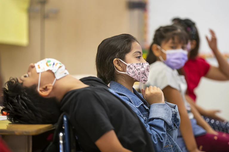 Four third-grade students wearing masks sit side by side in a classroom