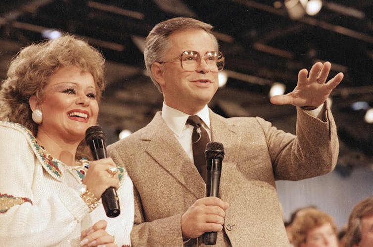 Man and woman hold microphones and speak before crowd.