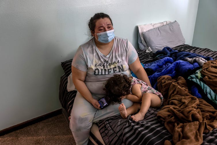 A young woman sitting on a bed with a small child who is hiding her face.