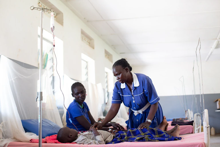 A female nurse standing over a child lying in a hospital bed. There's another woman seated next to the bed.