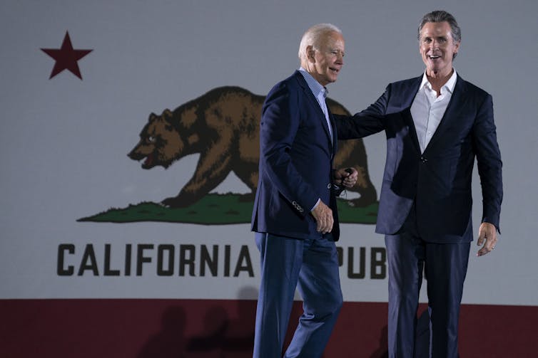 President Joe Biden greets California Gov. Gavin Newsom in front of a large California flag.