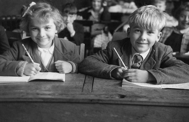 A boy and girl in 1950s school uniform sit at their desks holding pencils and smiling