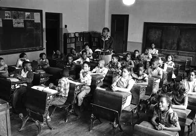 A black and white photo shows a teacher instructing a classroom of Black students.