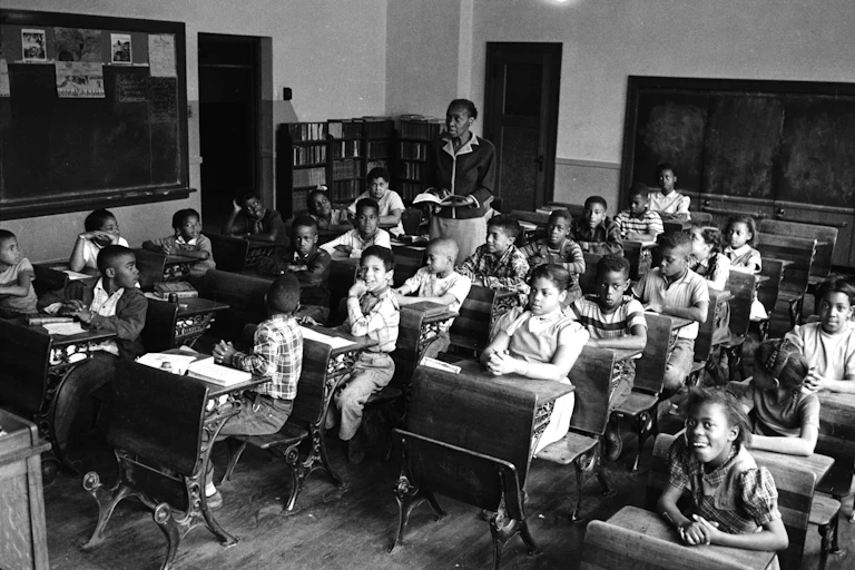 A black and white photo shows a teacher instructing a classroom of Black students.