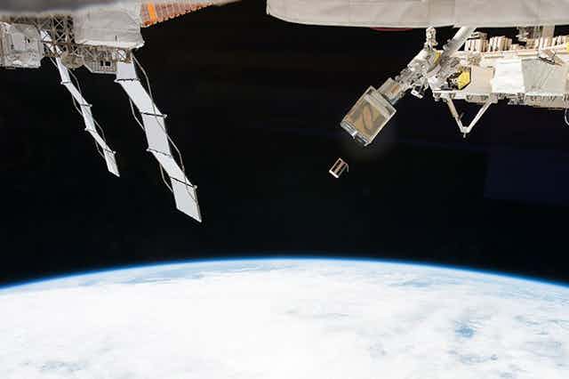 A small rectangular satellite drifting away from a hatch on the International Space Station.