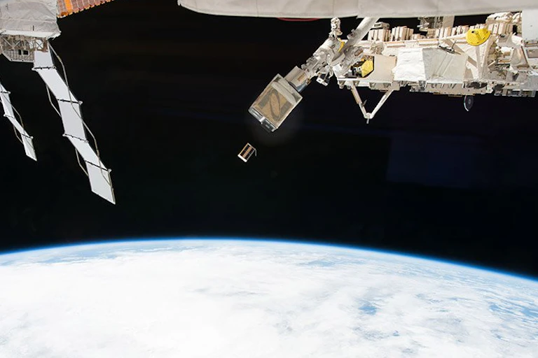 A small rectangular satellite drifting away from a hatch on the International Space Station.
