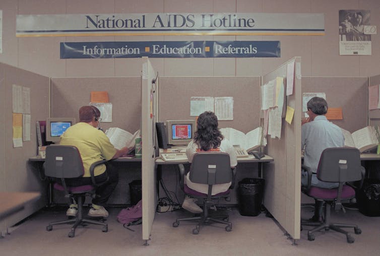 Three people at cubicle desks under a banner reading 'National AIDS Hotline - Information, Education, Referrals.'