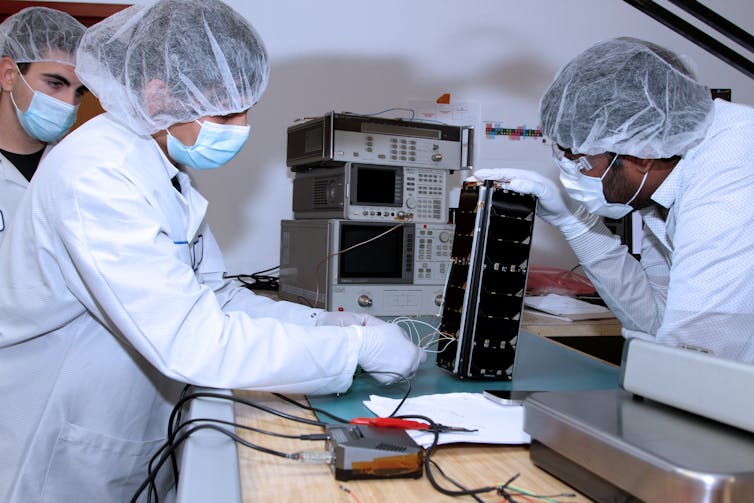 Three people in white lab coats and hairnets                      working on a satellite roughly the size of a loaf                      of bread.