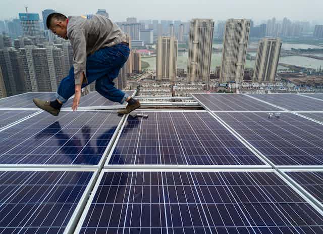 A man hunches over as he walks across solar panels on a skyscraper rooftop with a city of tall buildings along a port in the background.