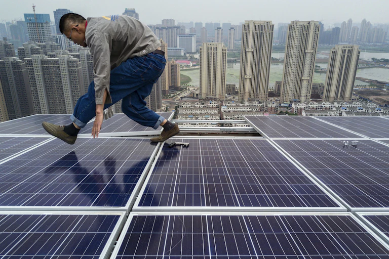 A man hunches over as he walks across solar panels on a skyscraper rooftop with a city of tall buildings along a port in the background.